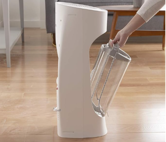 Honeywell Cool Moisture Tower Humidifier in a living room setting, showing a hand removing the easy-to-fill water tank from the side of the unit