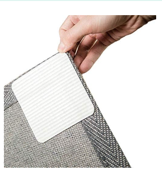 Hand lifting the corner of a grey woven rug to reveal a white, textured non-slip gripper pad attached to the underside.