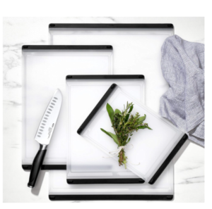 Set of three white OXO Good Grips cutting boards with black tapered grips, displaying a chef's knife and fresh herbs.