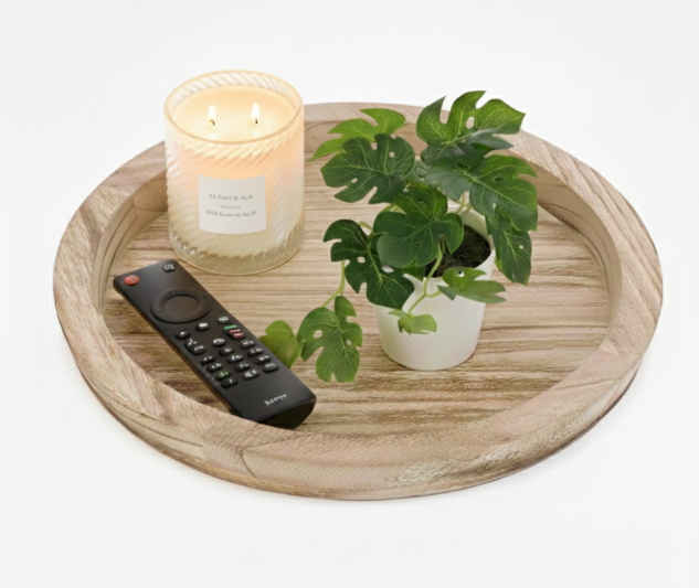 A round natural wood tray with a raised edge, styled as a coffee table centerpiece with a small potted plant, a lit candle, and a remote control on a white background.