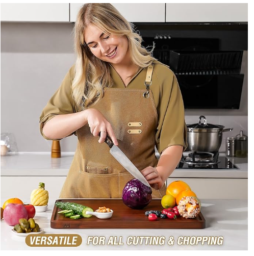 Smiling woman in an apron chopping red cabbage on a large, dark wood cutting board in a modern kitchen, with fresh vegetables and fruits nearby.