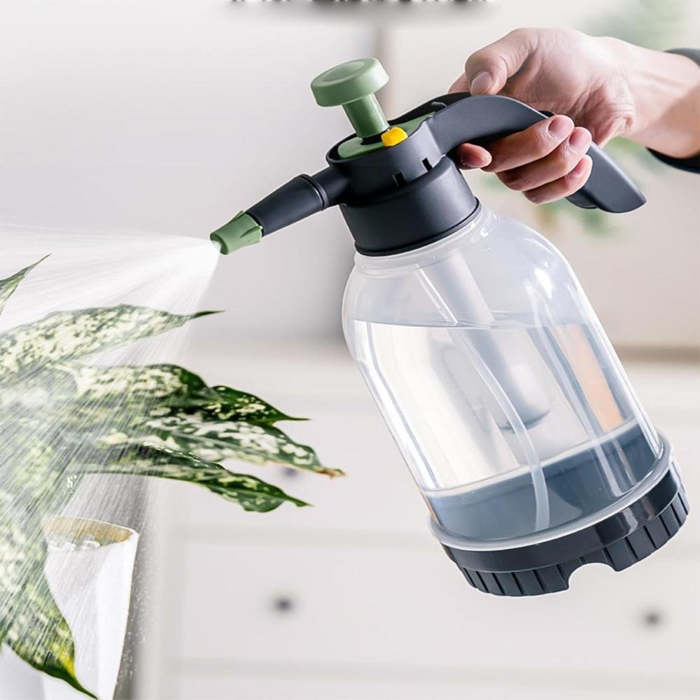 Hand holding large clear plastic pump-pressure plant sprayer with black base and green nozzle misting variegated plant leaves